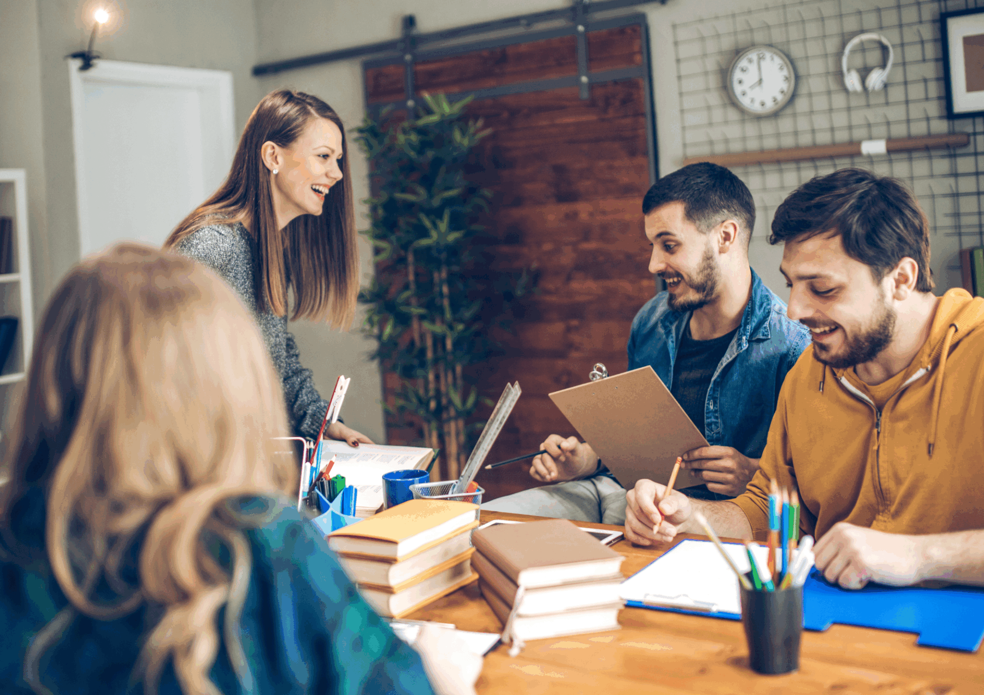 Fyra unga vuxna sitter runt ett bord och samarbetar i en avslappnad kontorsmiljö. De skrattar och diskuterar anteckningar med böcker och papper framför sig.