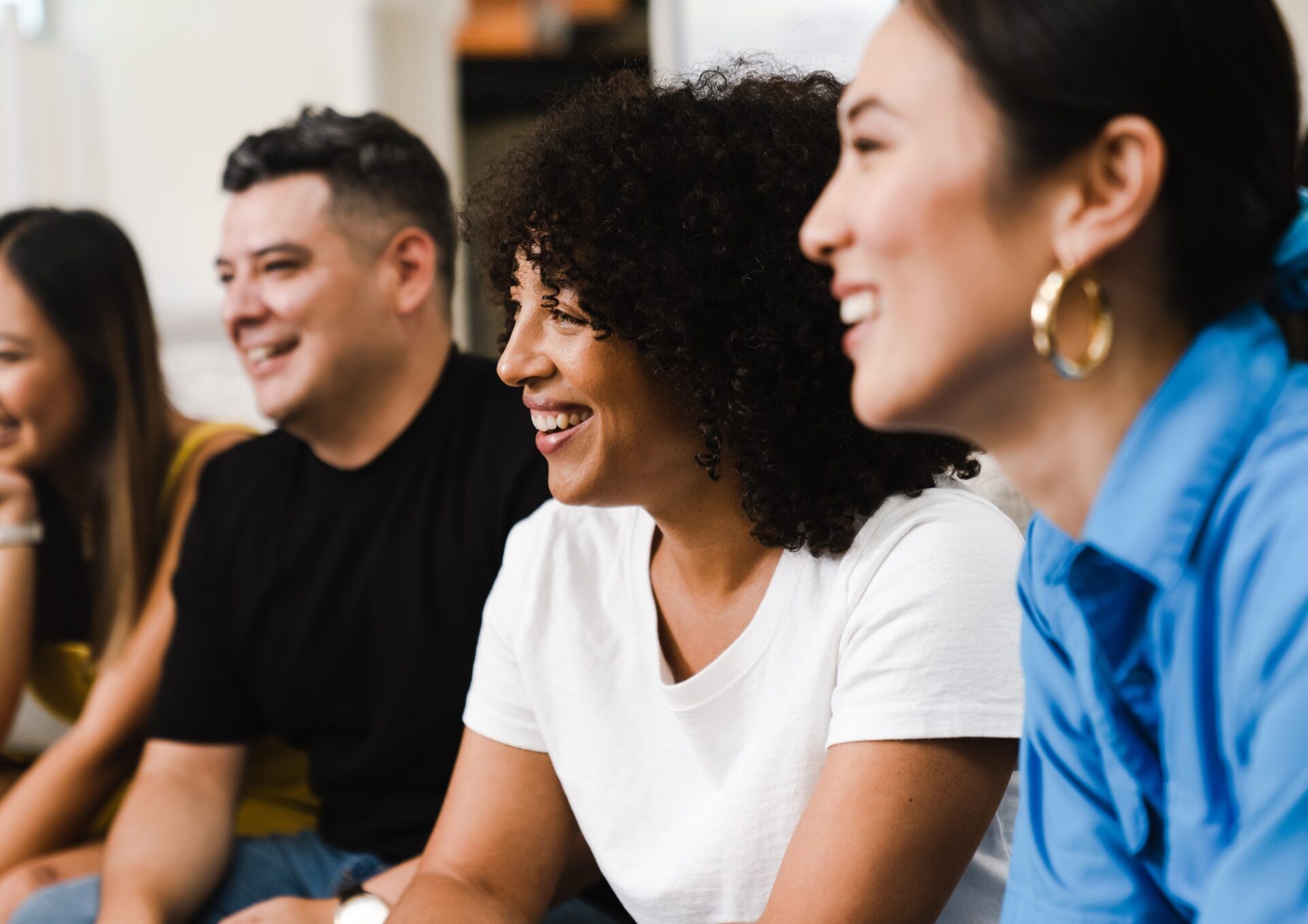 En grupp på fyra vuxna, med olika bakgrund, sitter i rad och ler under ett möte eller en aktivitet. Fokus ligger på de tre personerna i mitten och till höger.