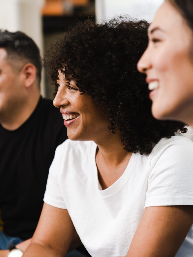 En grupp på fyra vuxna, med olika bakgrund, sitter i rad och ler under ett möte eller en aktivitet. Fokus ligger på de tre personerna i mitten och till höger.